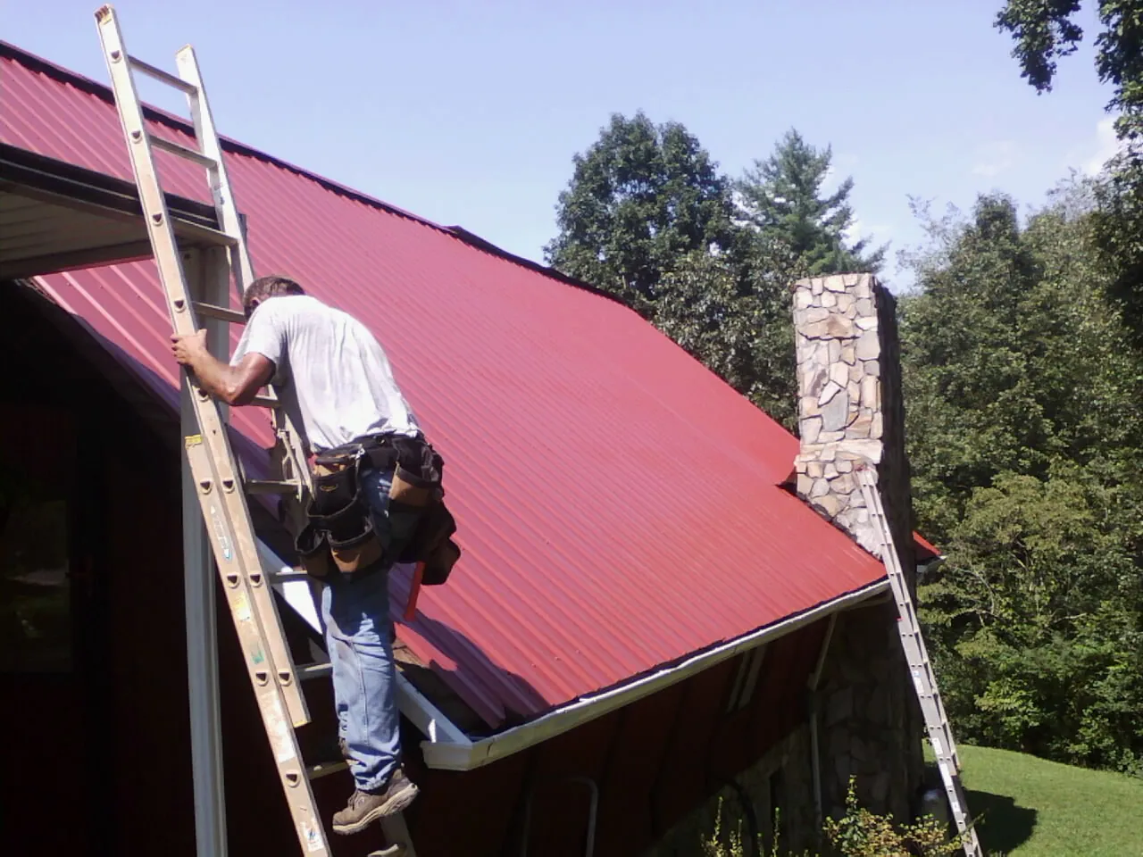 Roofer on a ladder beside a red metal roof and stone chimney — Soffit & Fascia Repair by Roofing Brothers in Reedley